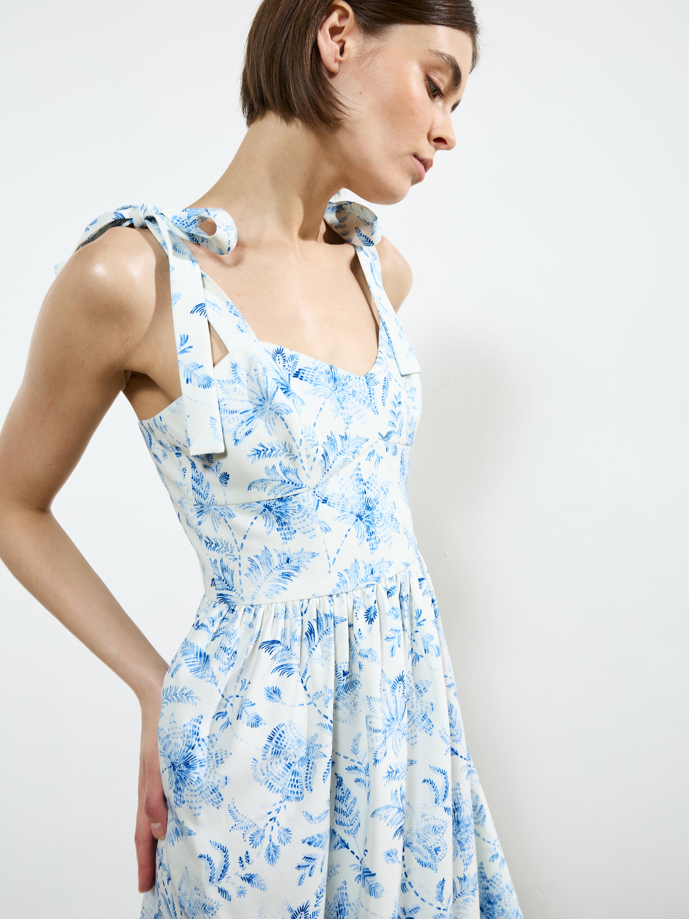 A woman wears the French Connection Serenity Dale Crepe Midi Dress, featuring a blue botanical print and tied shoulder straps, as she stands against a plain white background and looks slightly downward.