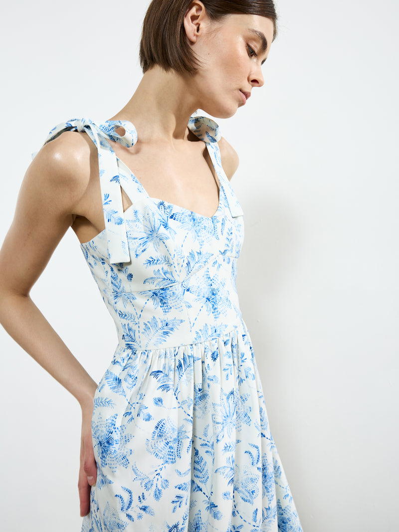 A woman wears the French Connection Serenity Dale Crepe Midi Dress, featuring a blue botanical print and tied shoulder straps, as she stands against a plain white background and looks slightly downward.