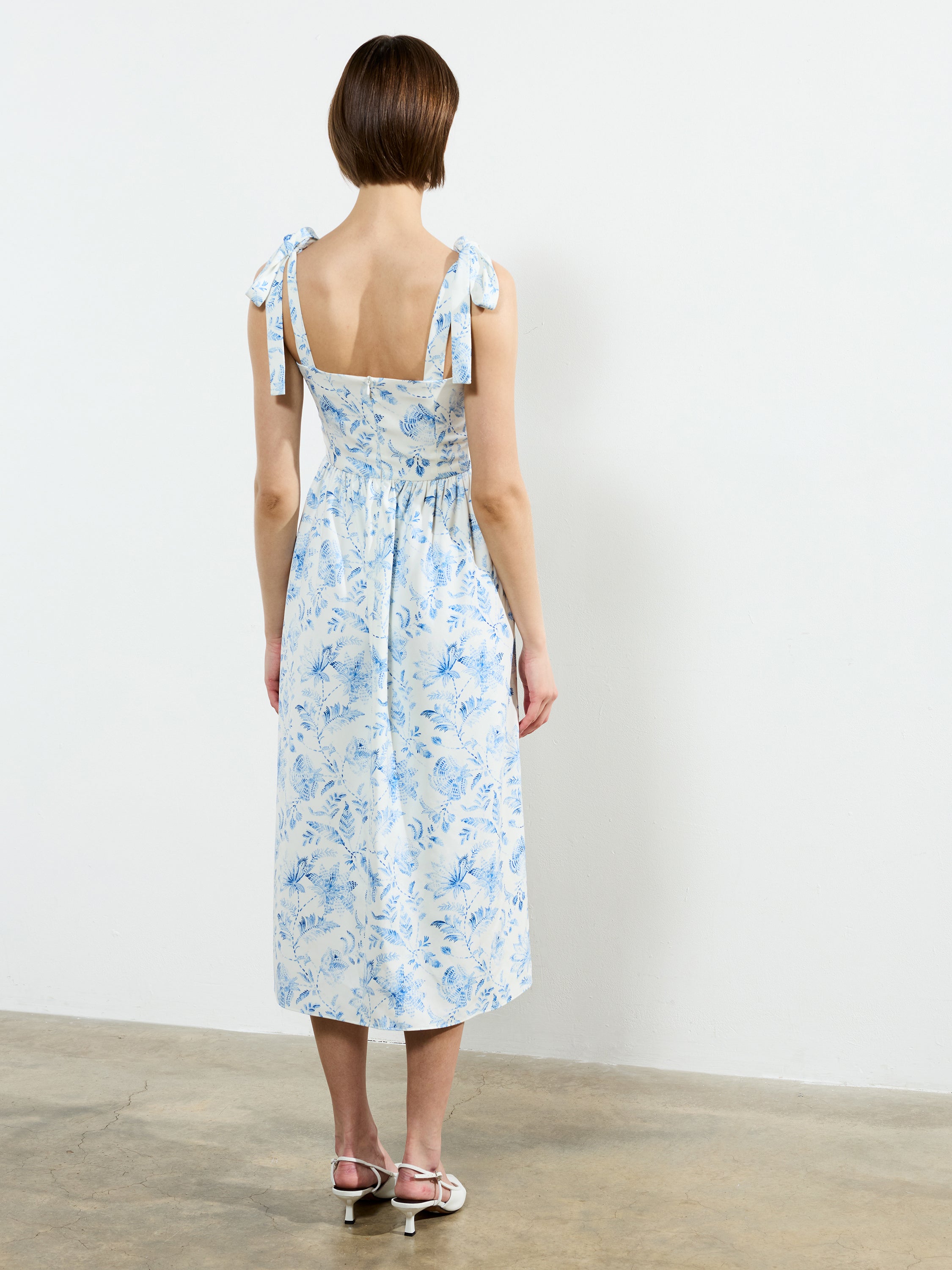 A woman with short brown hair, seen from behind, wears the French Connection Serenity Dale Crepe Midi Dress—white with a blue floral print and bow-tied straps—standing on a concrete floor against a white wall.