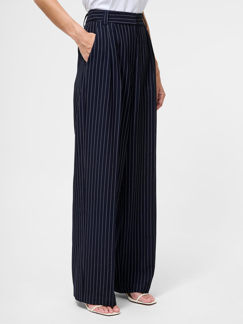 A person wears the Pinstripe Tailored Wide Leg Trousers in navy with white stripes, paired with a tucked-in white top and open-toe heels, standing hands in pockets against a plain background.