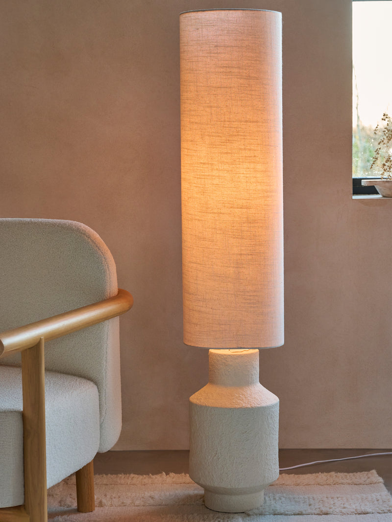 Tall floor lamp with a textured shade next to a chair in a room with a window.