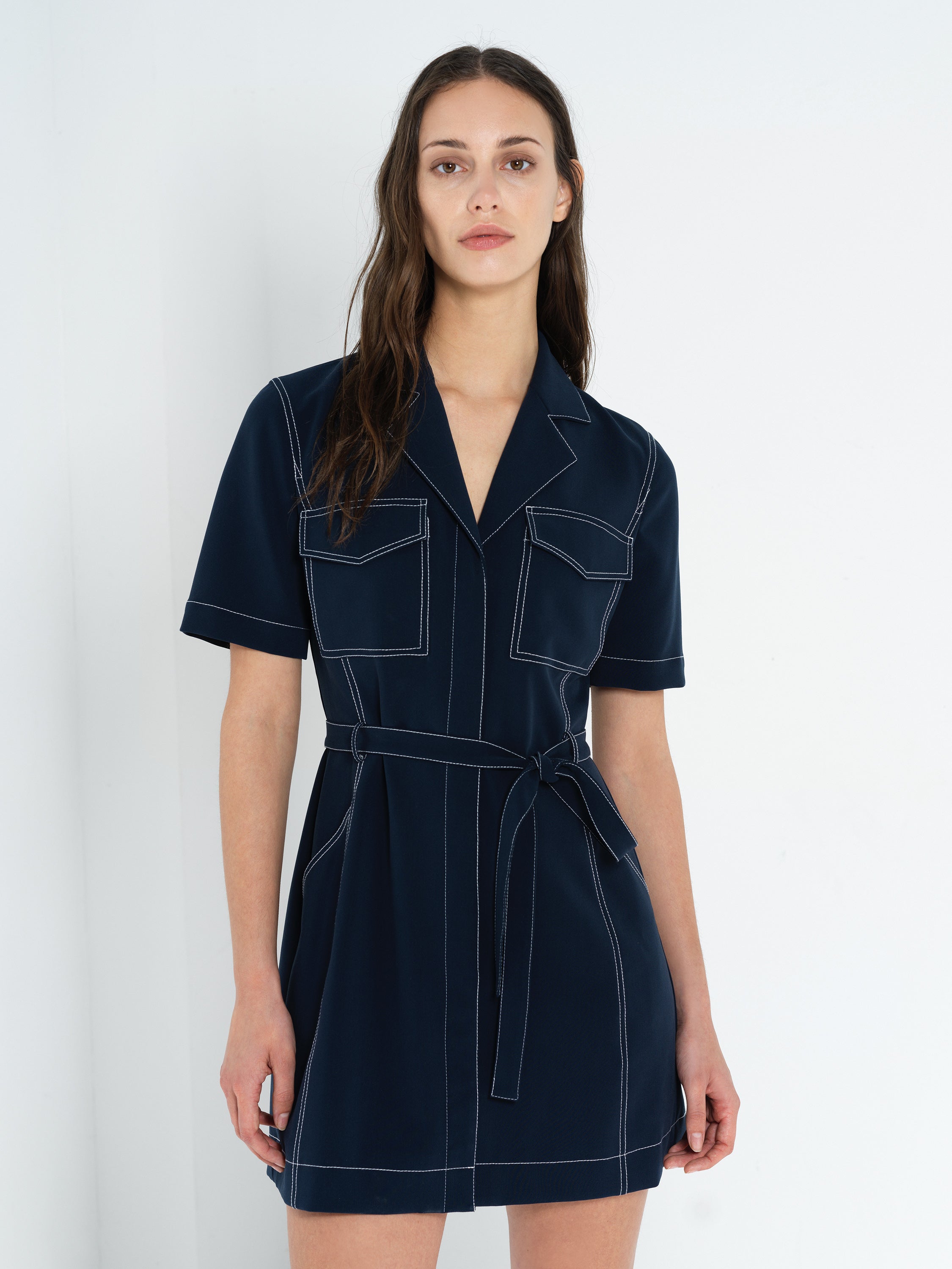 A woman wearing a belted navy blue mini dress with contrast stitching, a revere collar and button down fastening.