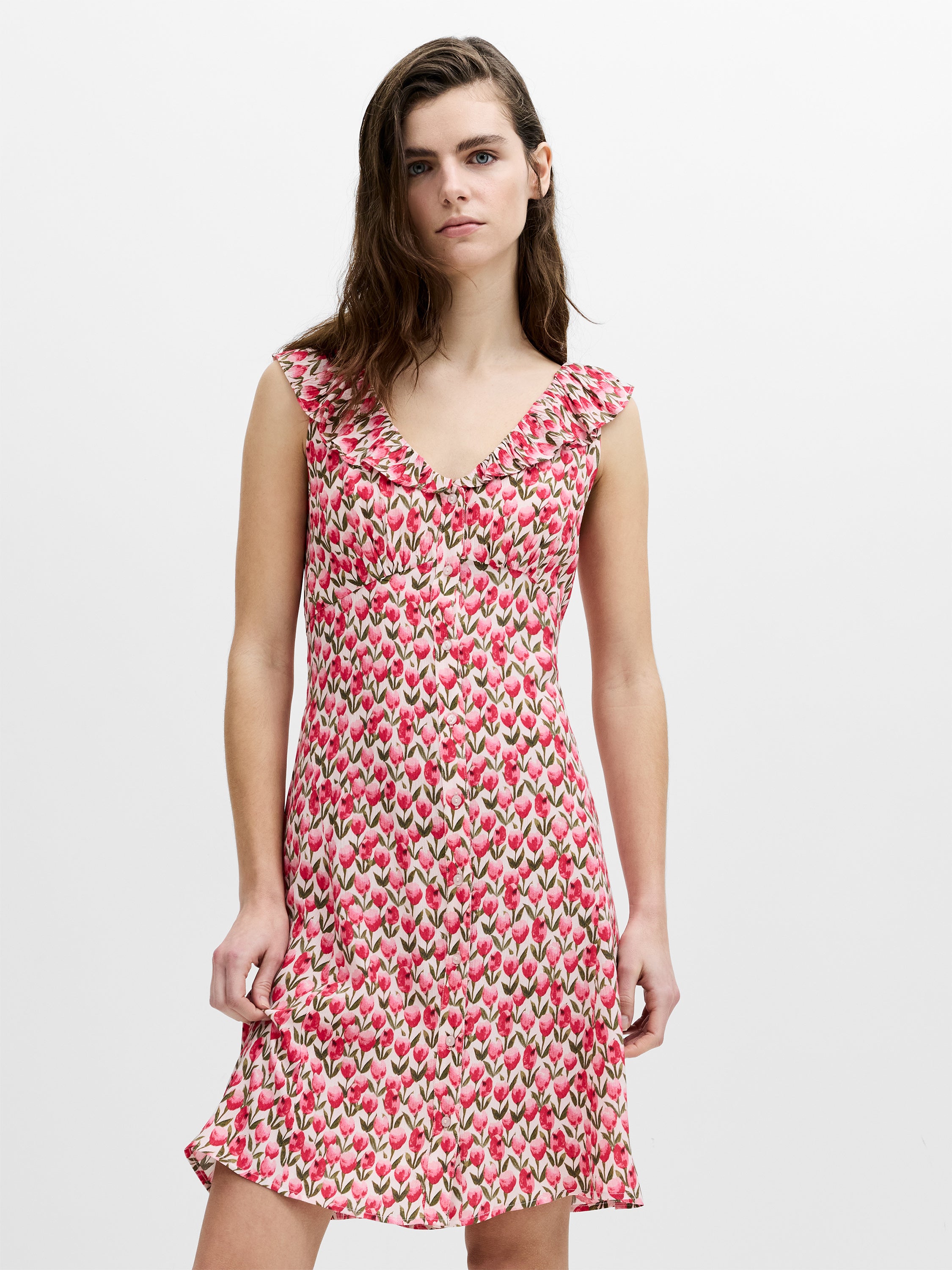 A woman wearing a pink tulip print mini dress with a V-neckline and frill trim, an empire waistline and button down fastening.