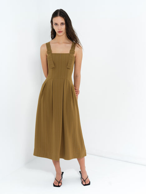 A woman wearing a khaki pinstripe buckle adjustable strap midi dress with a square neckline and side pockets, a drop waist and pleated skirt.