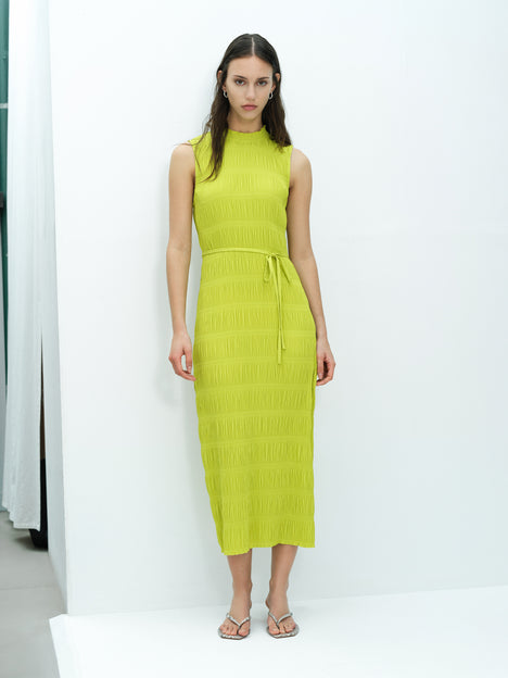 A woman wearing a sleeveless green midi length dress with a tie waist, high frill neckline and side split hem.