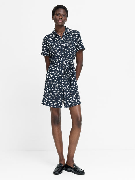 A woman wearing a short sleeve mini length navy dress with a white floral print, shirt collar, self tie belt and a button down fastening. 