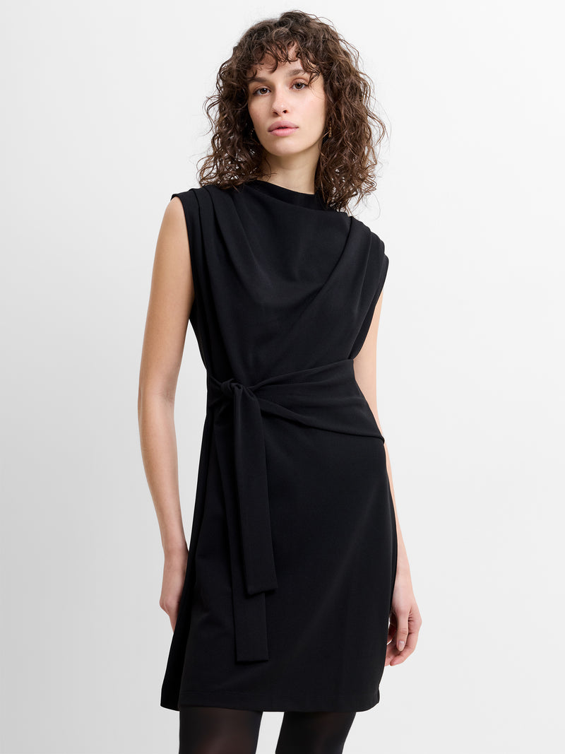 Woman wearing a sleeveless black belted mini dress with a draped high neckline and a self tie waist belt