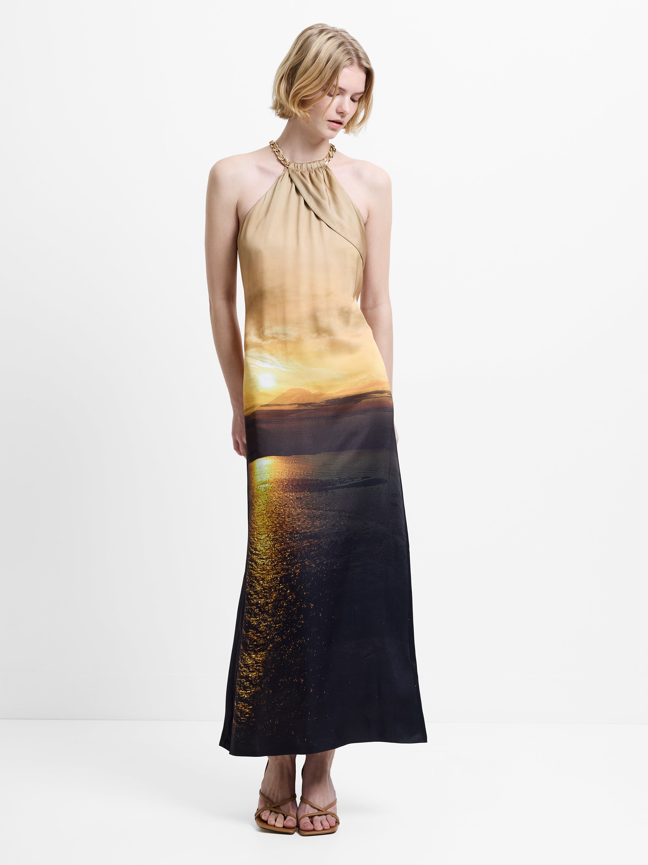 Woman wearing a halterneck, satin midi dress with a scenic sunset print.