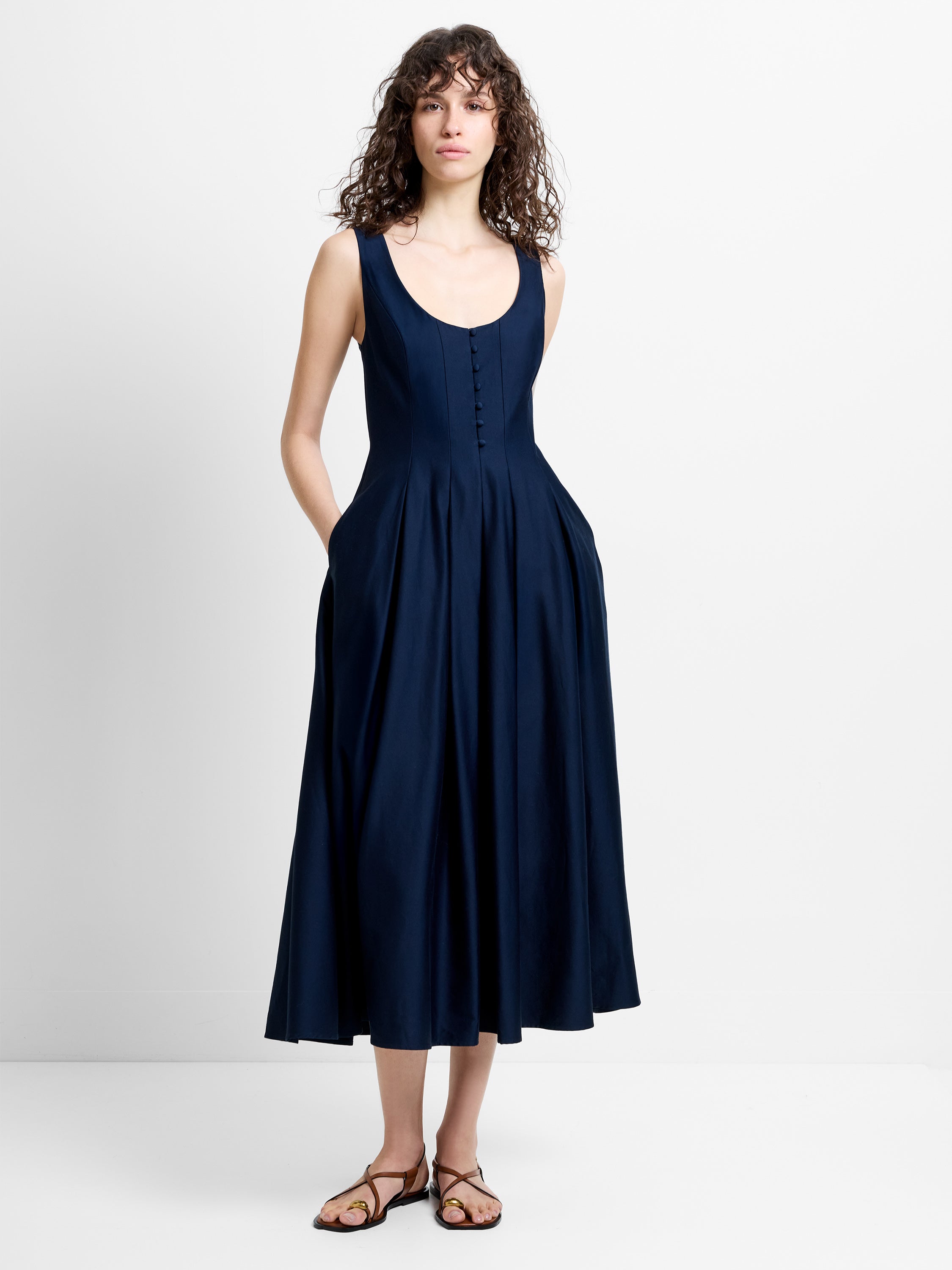 Woman wearing a navy blue sateen midi dress, with a deep scoop neckline, straps and a flared pleated skirt