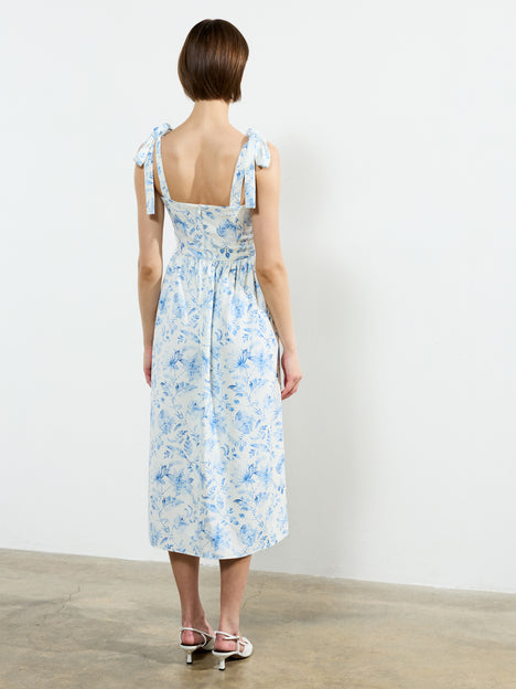 A woman with short brown hair, seen from behind, wears the French Connection Serenity Dale Crepe Midi Dress—white with a blue floral print and bow-tied straps—standing on a concrete floor against a white wall.