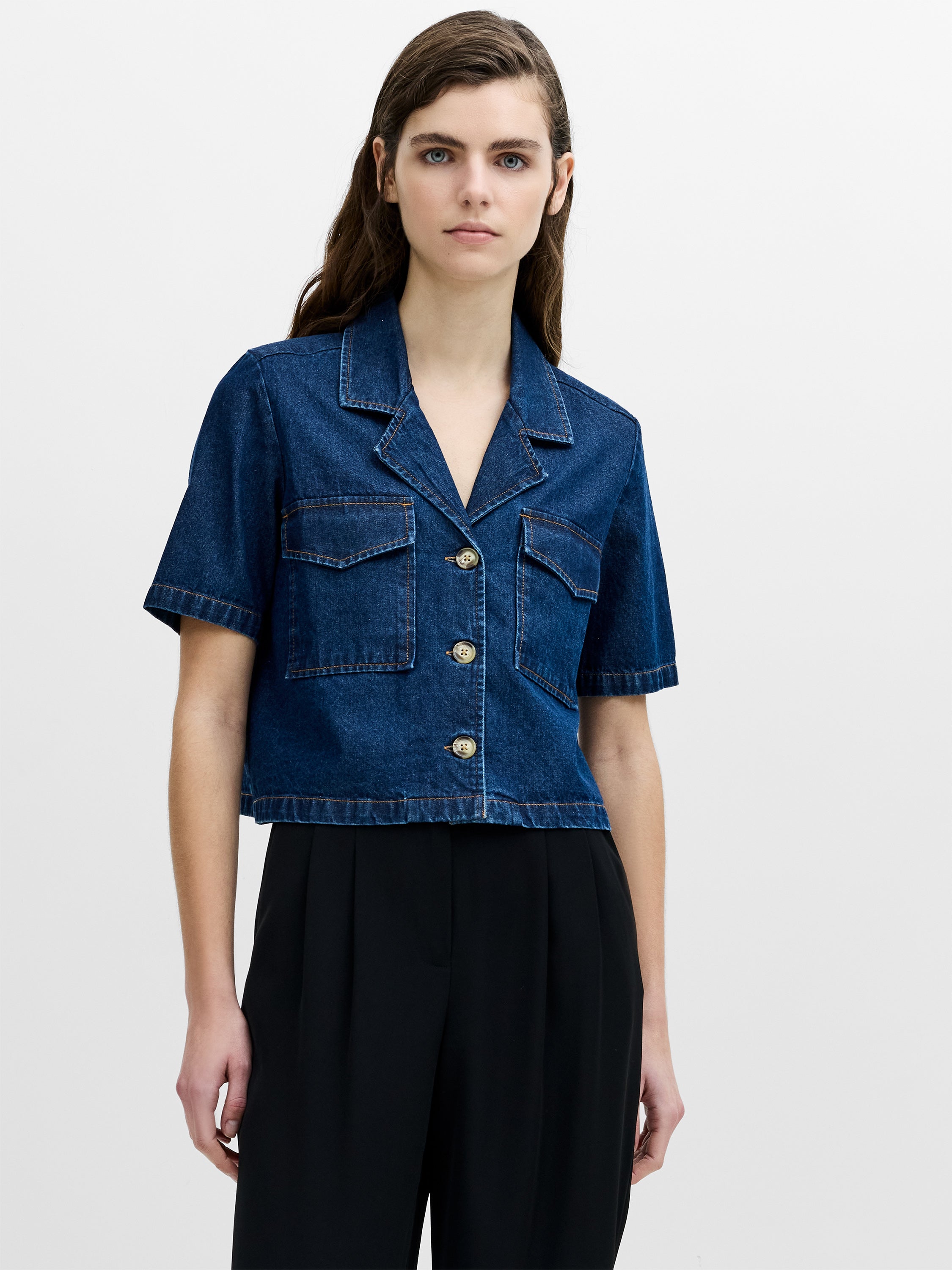 A woman wearing a short sleeved blue denim shirt with a revere collar, chest pockets, a boxy cropped fit and a button through fastening.