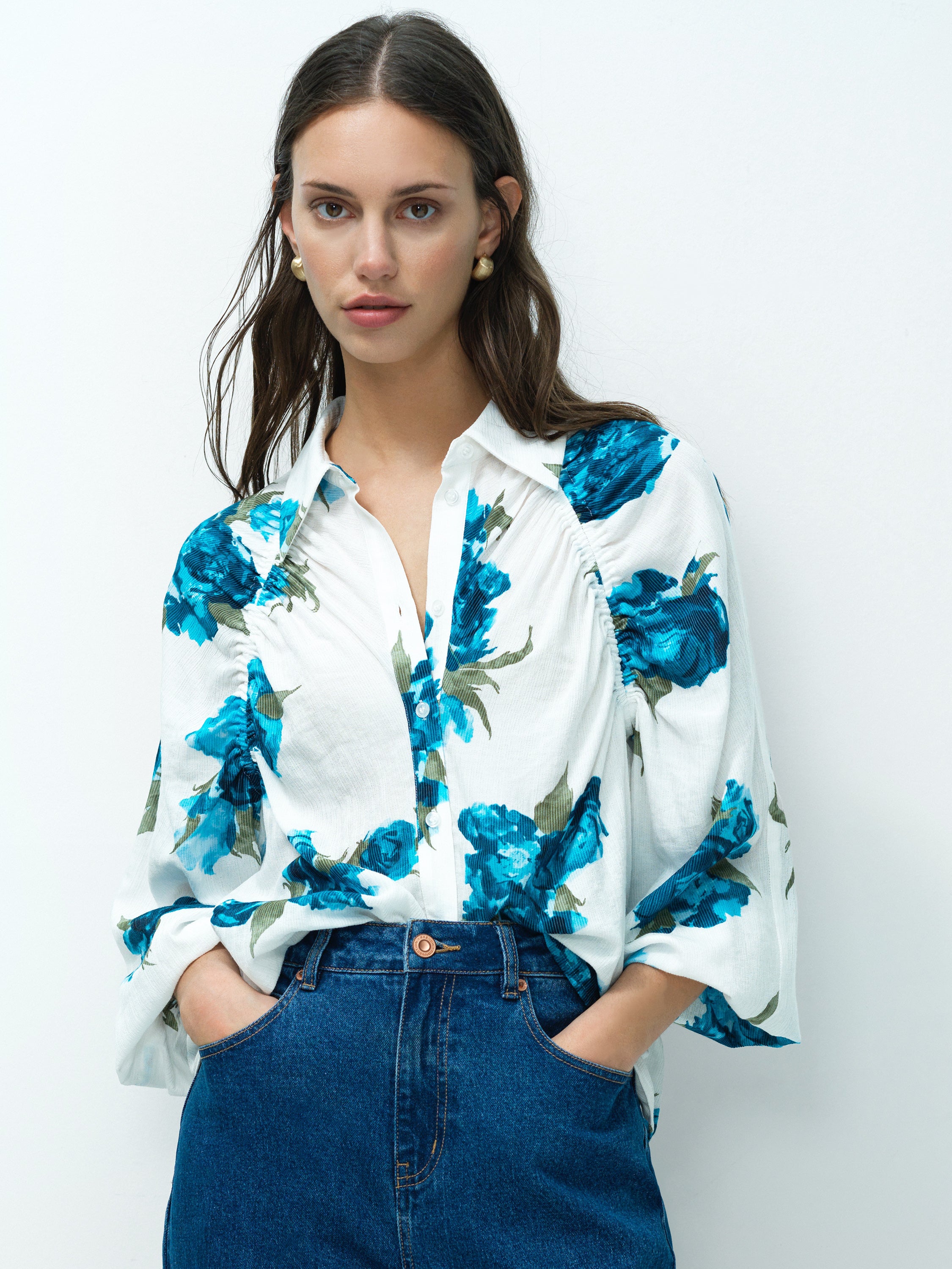 Woman wearing a point collar white blouse with blue floral print and long blouson sleeves and a button down fastening. 