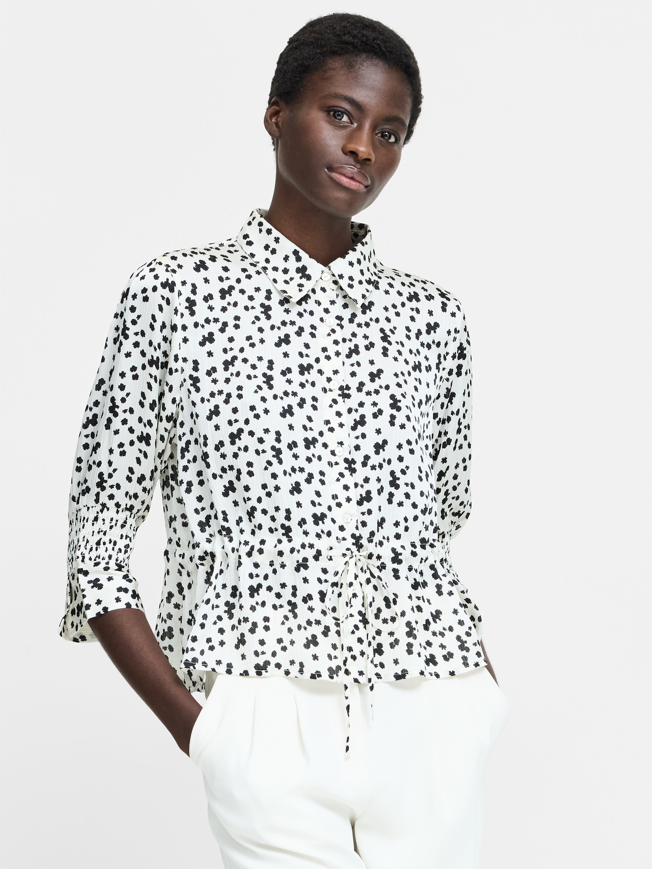 A woman wearing a cream button down shirt with a black floral pattern, 3/4 length sleeves, a collared neckline, and a self tie drawstring waist.