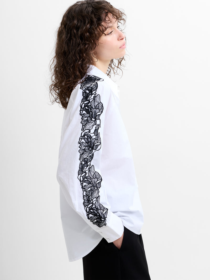 Woman wearing a white long sleeve shirt, with black floral embroidered sleeve detailing 