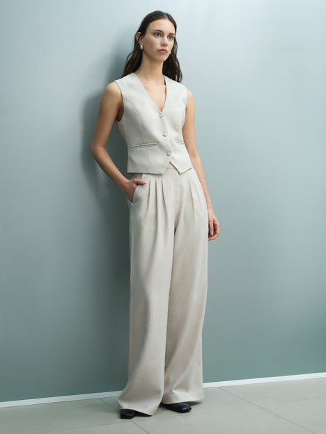 A woman wearing tailored high waisted, full length pleated, wide leg, beige trousers with four pockets.