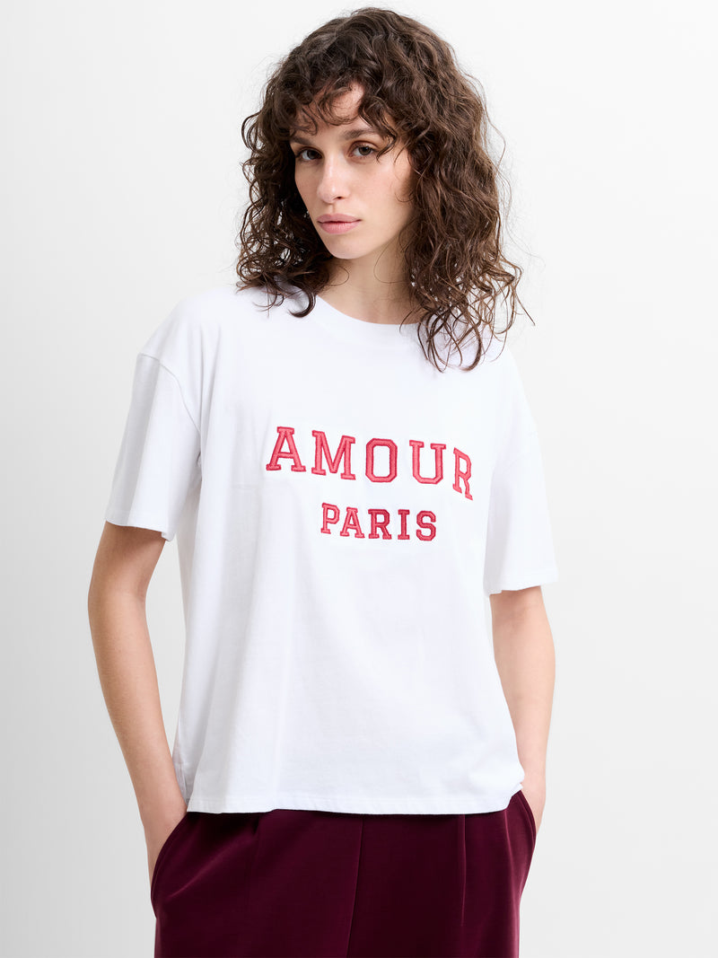 Person wearing a white short sleeve relaxed fit T-shirt with 'AMOUR PARIS' graphic