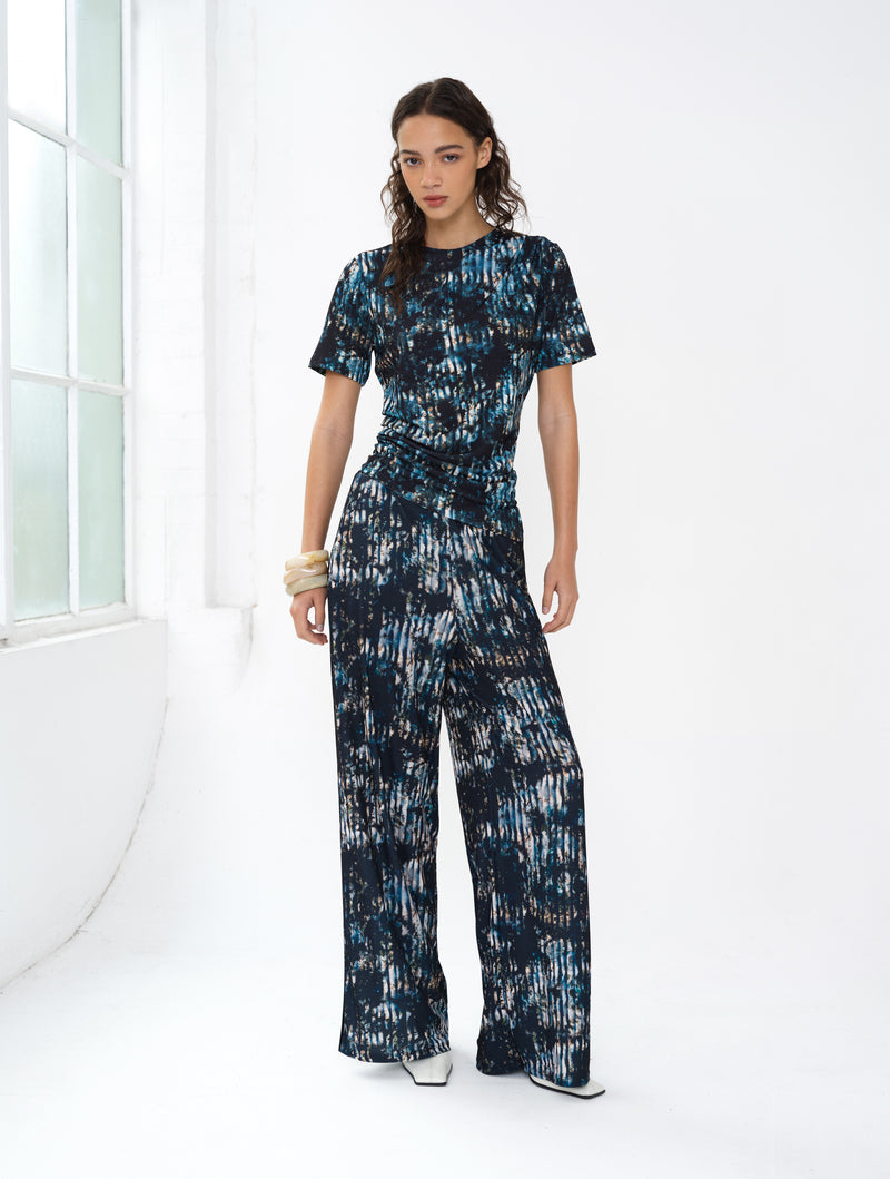 Woman wearing an abstract printed co-ord. A short sleeve draped top and wide leg trousers.