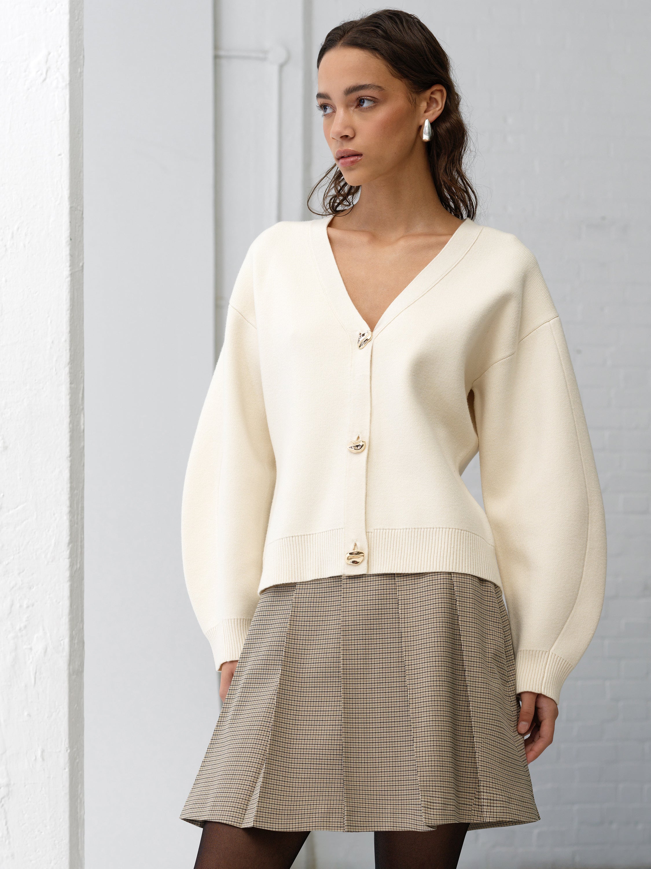 Woman wearing a cream long sleeve cardigan with a cinched waist, with a check pleated mini skirt