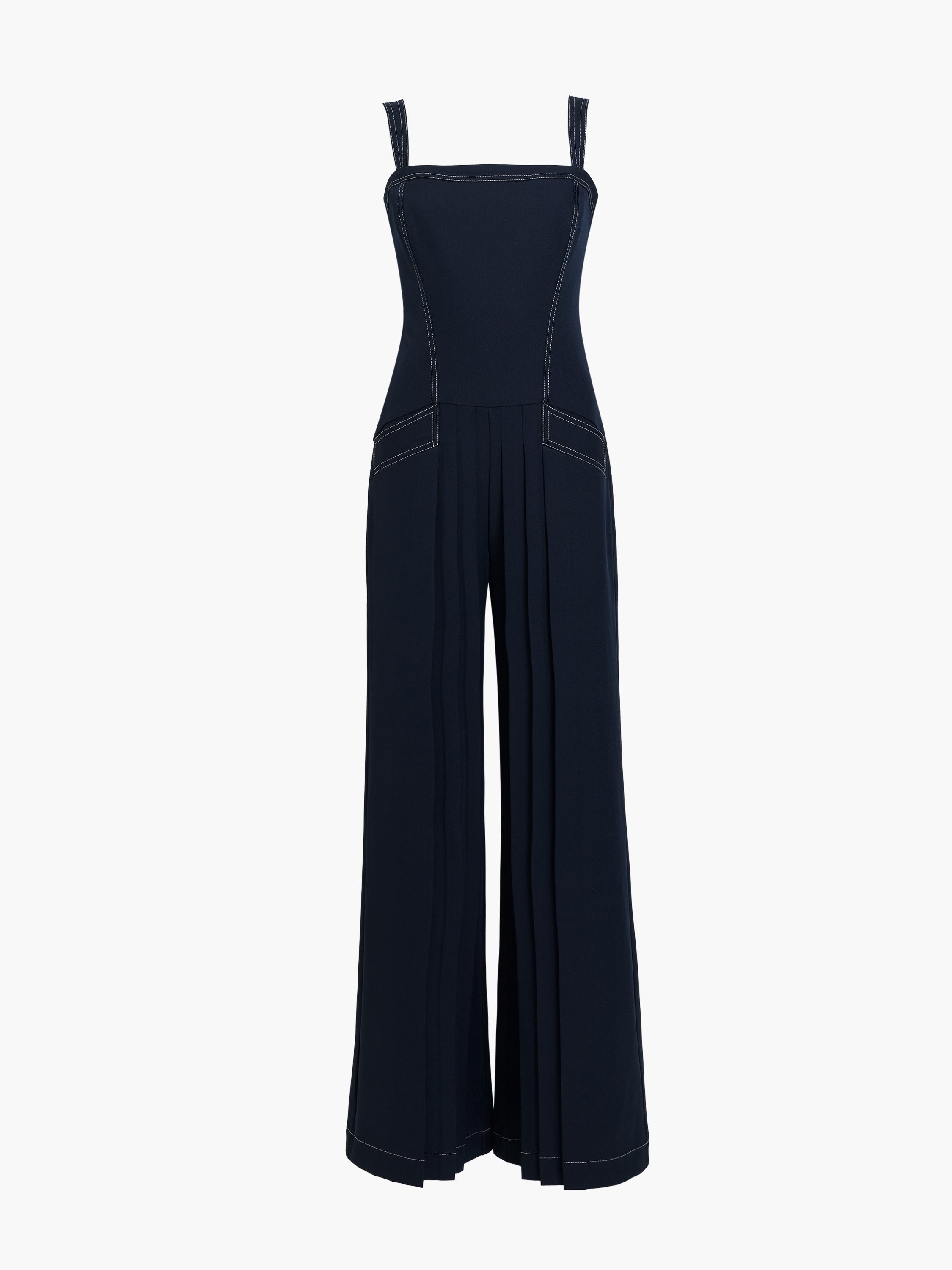 Woman wearing a pleated navy blue full length wide leg jumpsuit with straps, a square neckline, front pockets, dropped waist and contrast stitching.