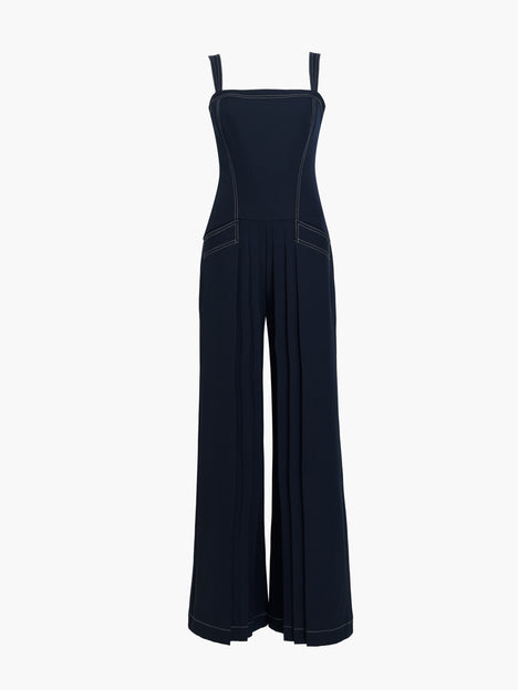 Woman wearing a pleated navy blue full length wide leg jumpsuit with straps, a square neckline, front pockets, dropped waist and contrast stitching.